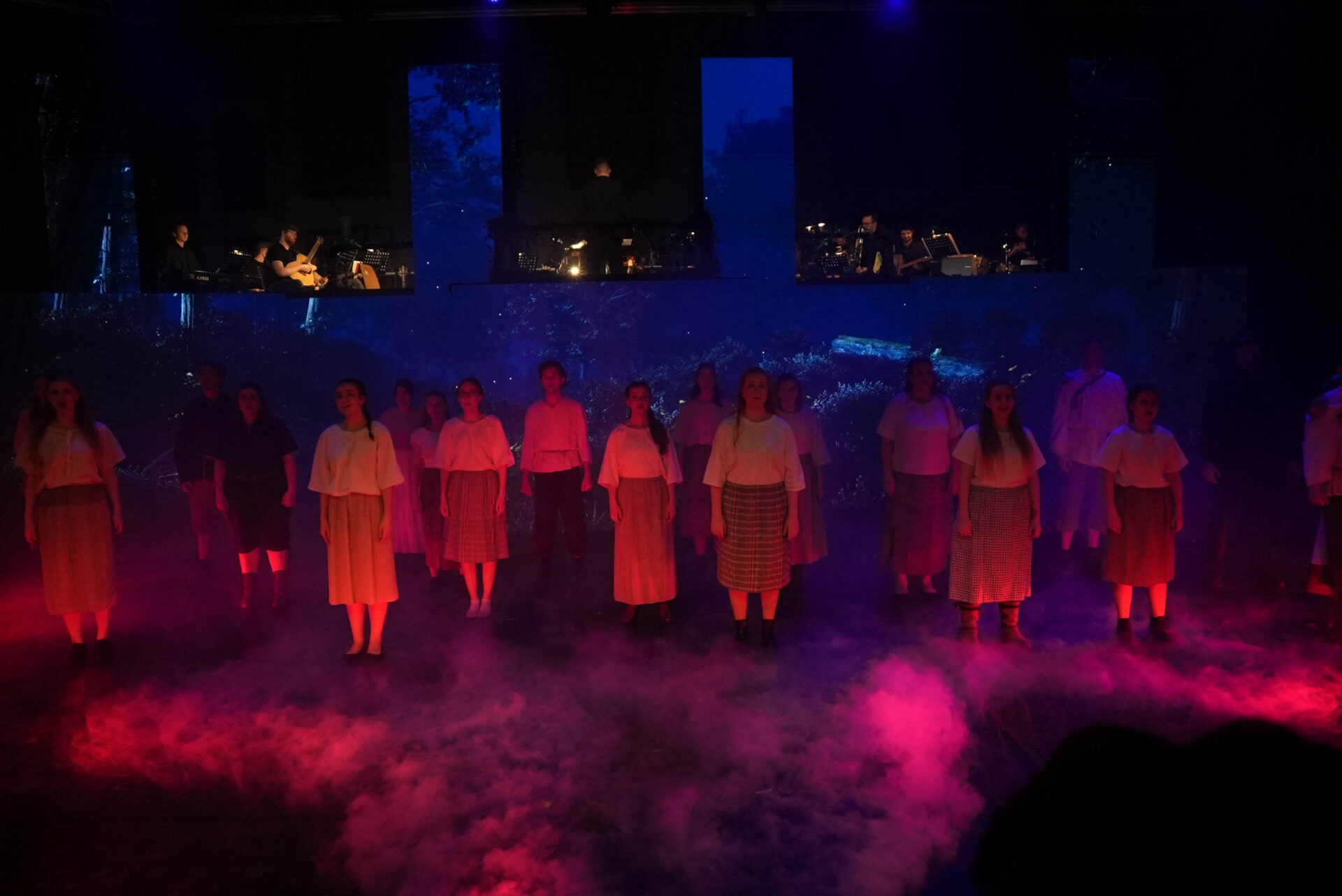 Das Ensemble steht in dichtem Bühnennebel unter rotem und blauem Licht auf der Bühne, während das Orchester erhöht im Hintergrund spielt.