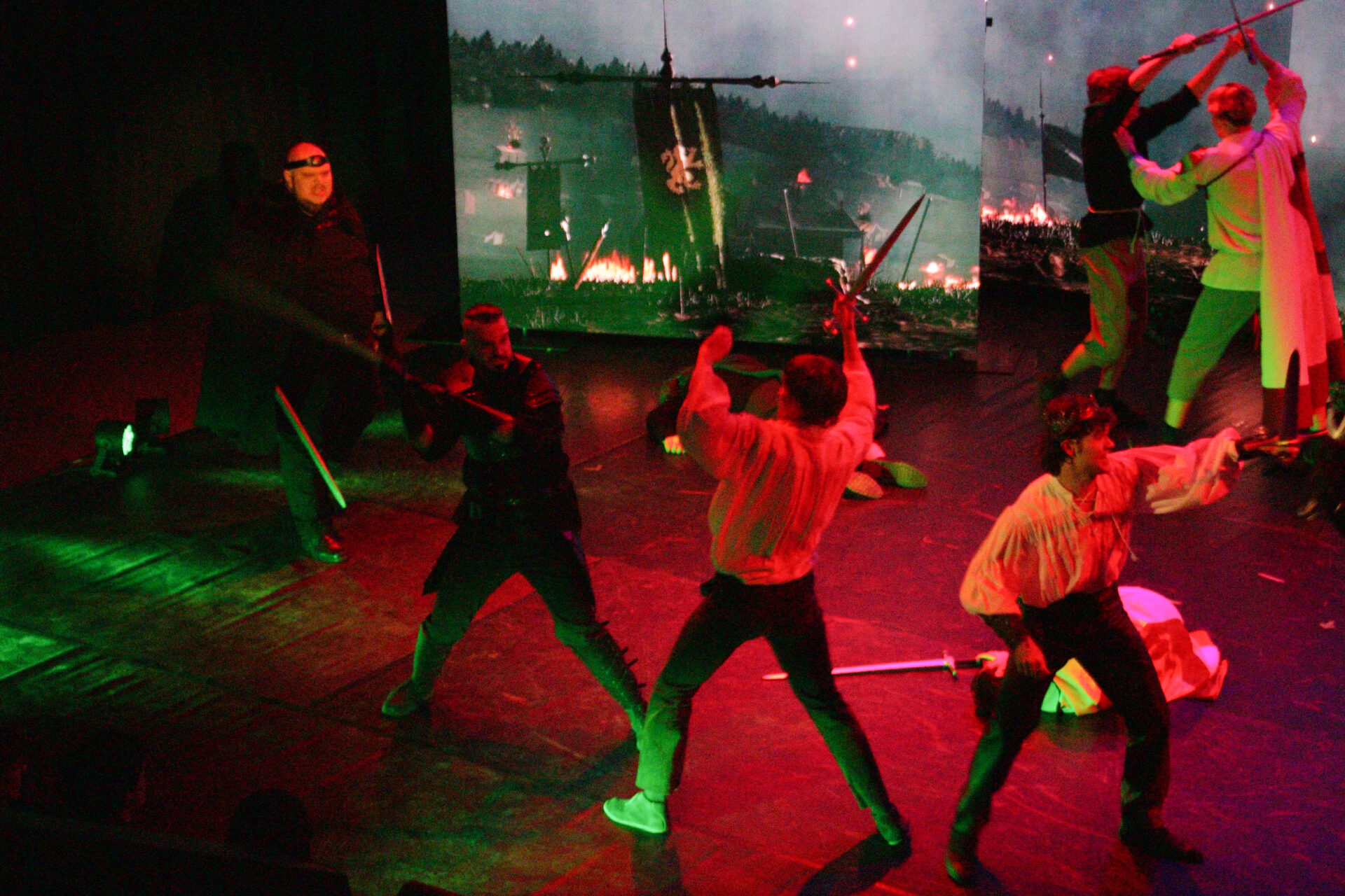 Mehrere Darstellende liefern sich auf einer Theaterbühne in rotem und grünem Licht eine Schwertkampfszene vor einer kriegerischen Projektion im Hintergrund.