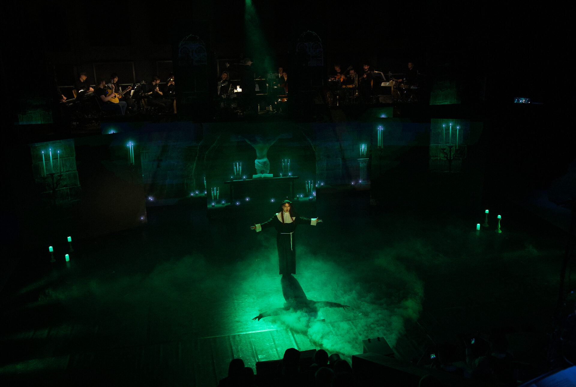 Eine Solistin steht in grünem Licht und Bühnennebel im Mittelpunkt der Szene, während das Orchester erhöht im Hintergrund über das Geschehen blickt.